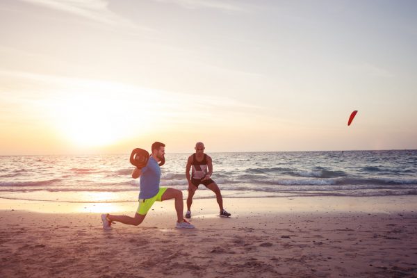 Man doing fitness on the beach with the help of personal trainer. Warm tone, lens flare.
